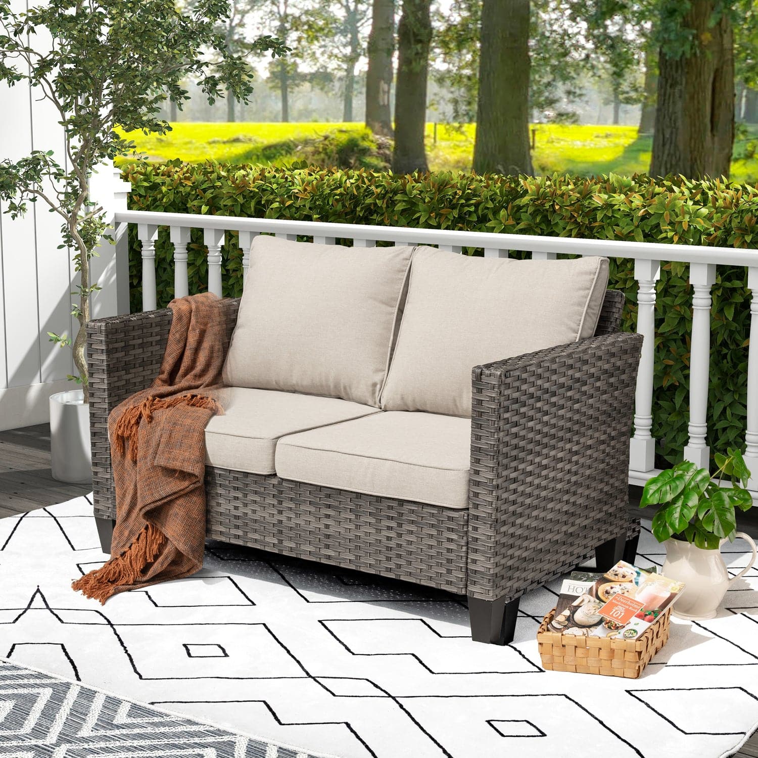 An Ovios grey wicker loveseat with beige cushions sits on a patterned rug, overlooking a lush green backyard