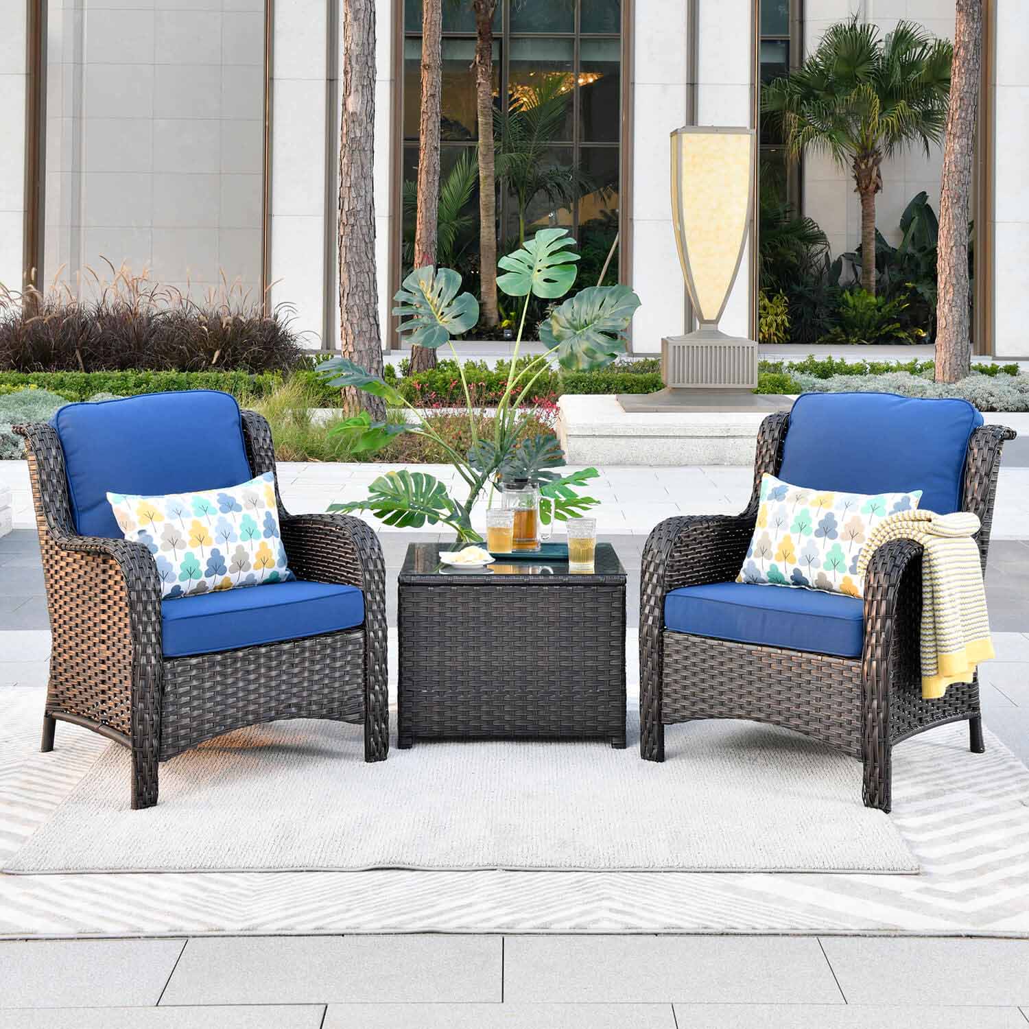 The Ovios dark wicker bistro set with vibrant blue cushions and patterned pillows creates a stylish seating area on the sunny patio