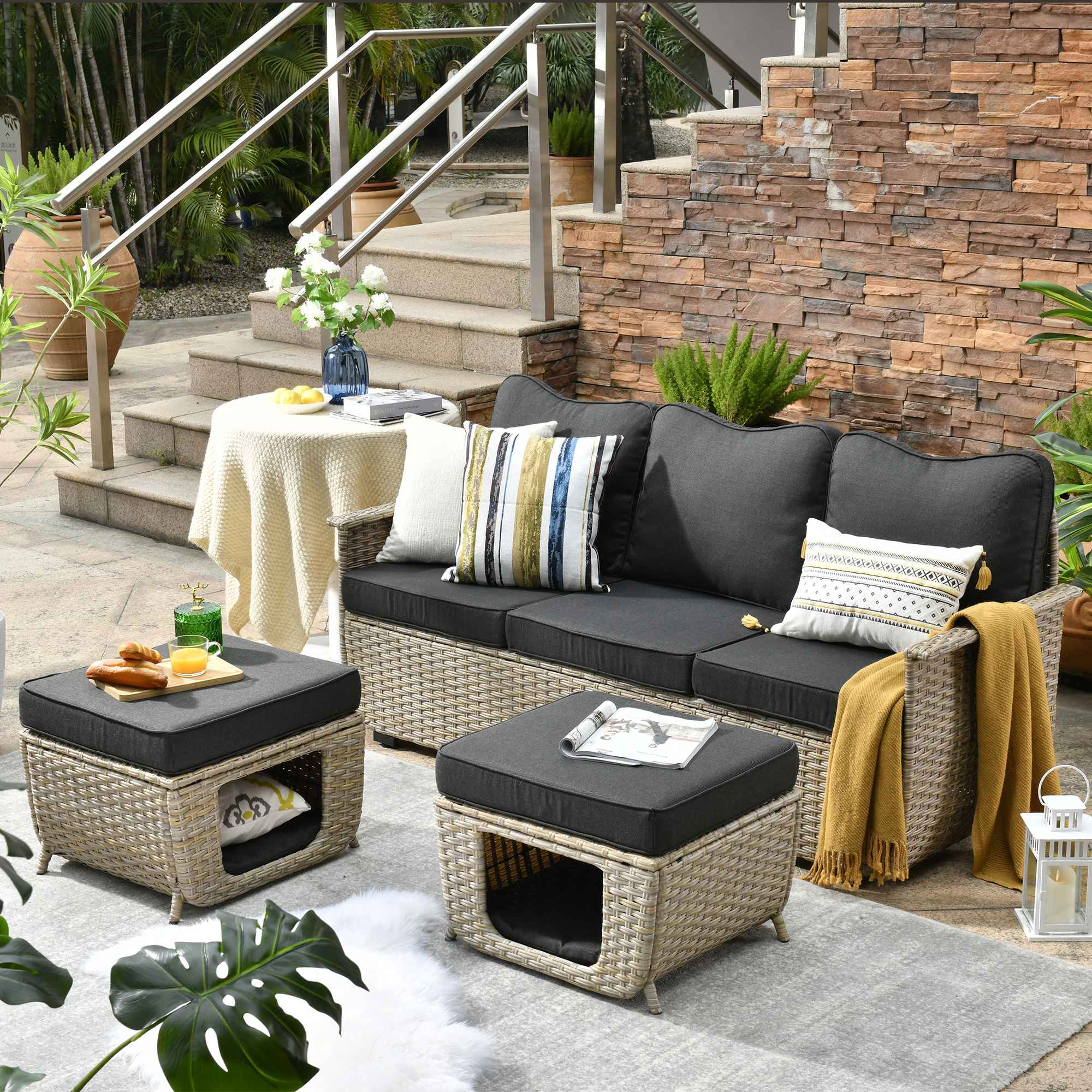 The Ovios beige wicker patio sofa with dark gray cushions and ottomans creates a cozy lounge on the stone terrace beside brick stairs