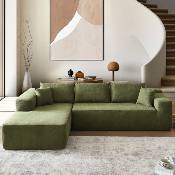 An Ovios olive green sectional sofa sits in a minimalist room with a spiral staircase and abstract wall art